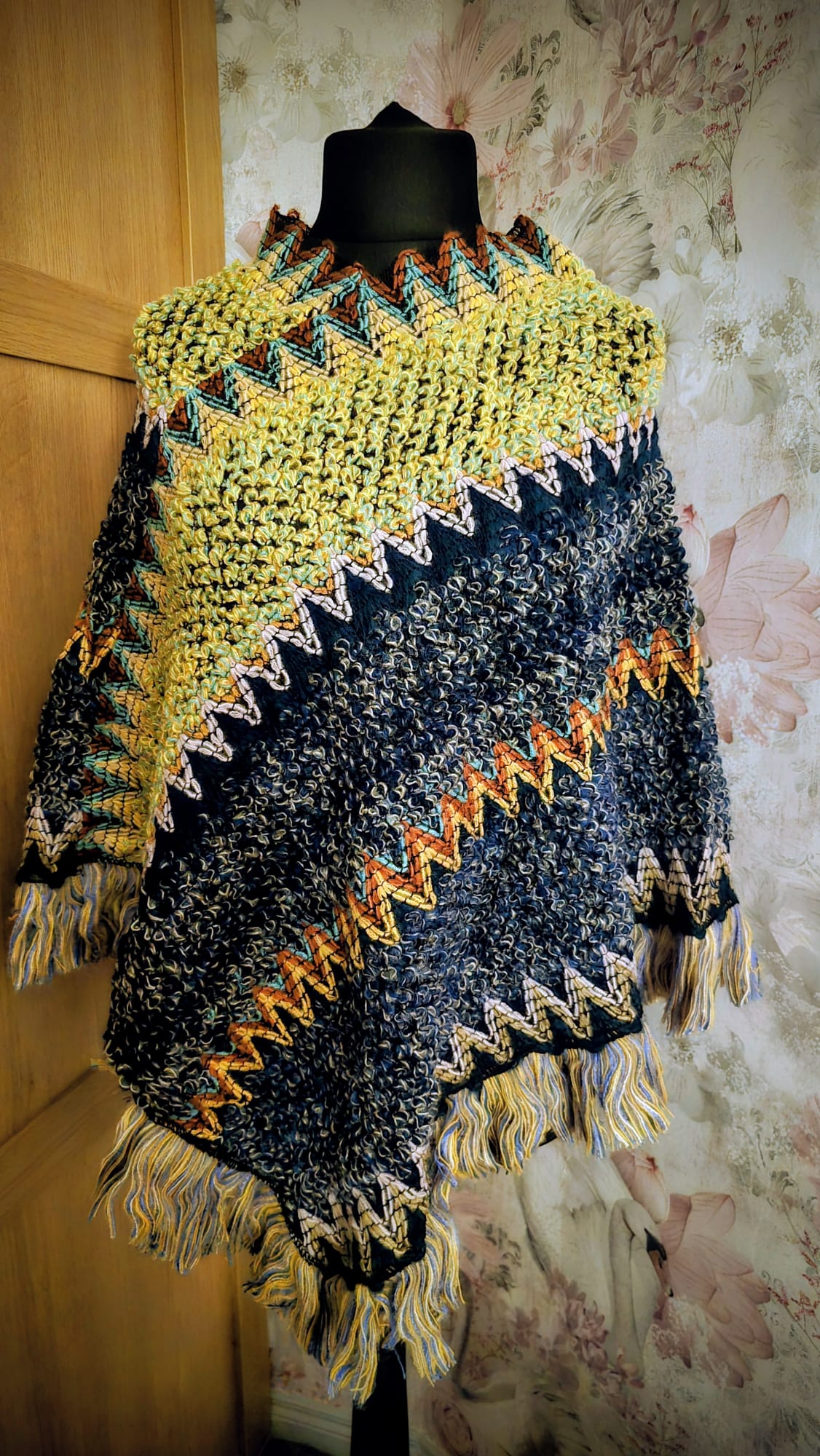 Colorful knitted poncho on a mannequin against a textured wall.