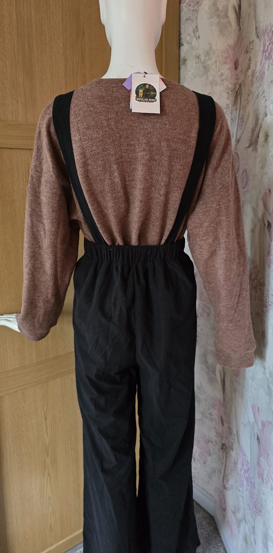 Mannequin wearing a brown sweater and black overalls with a tag, against a wooden door and floral wall.