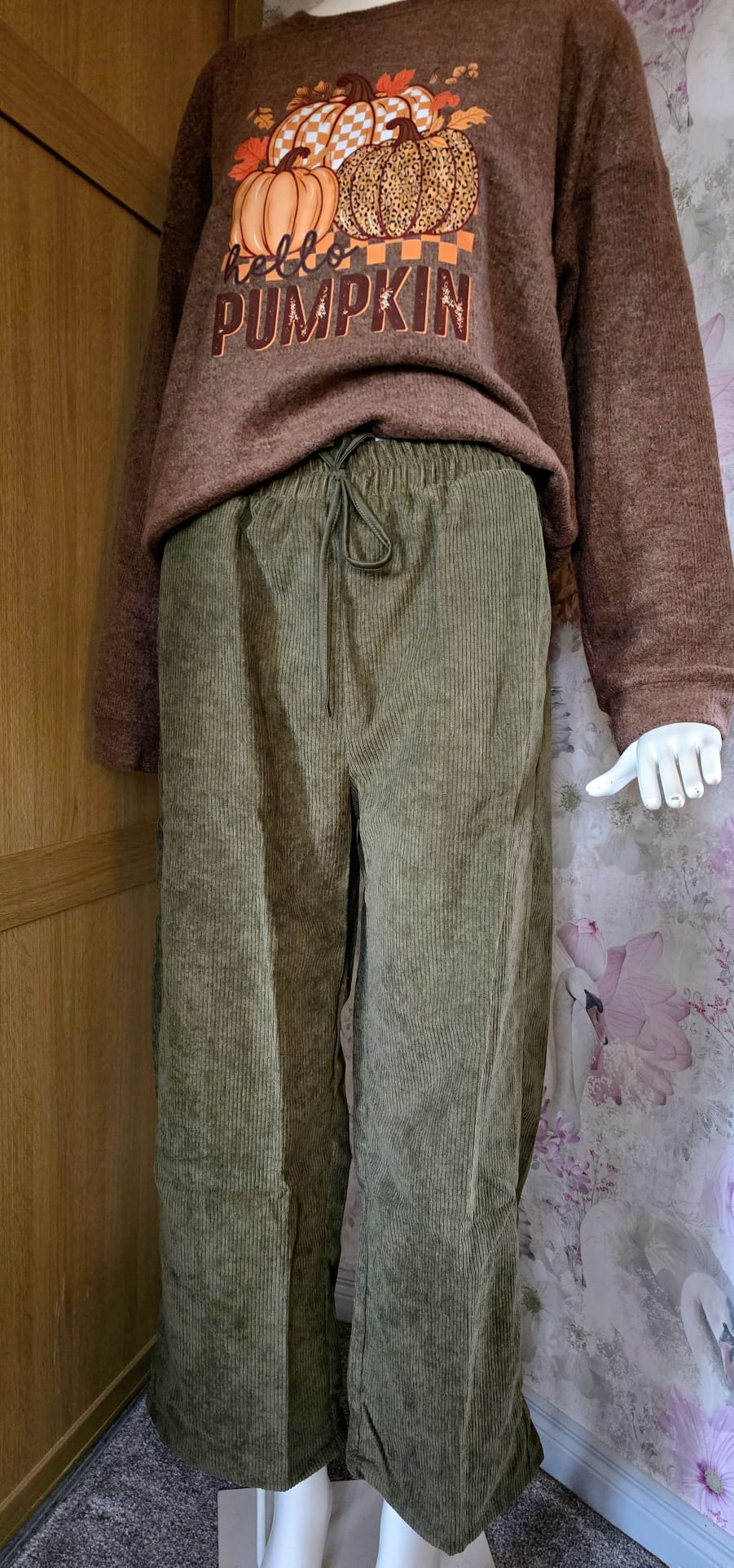 Brown sweater with pumpkin design and green pants on a mannequin against a floral wallpapered wall.